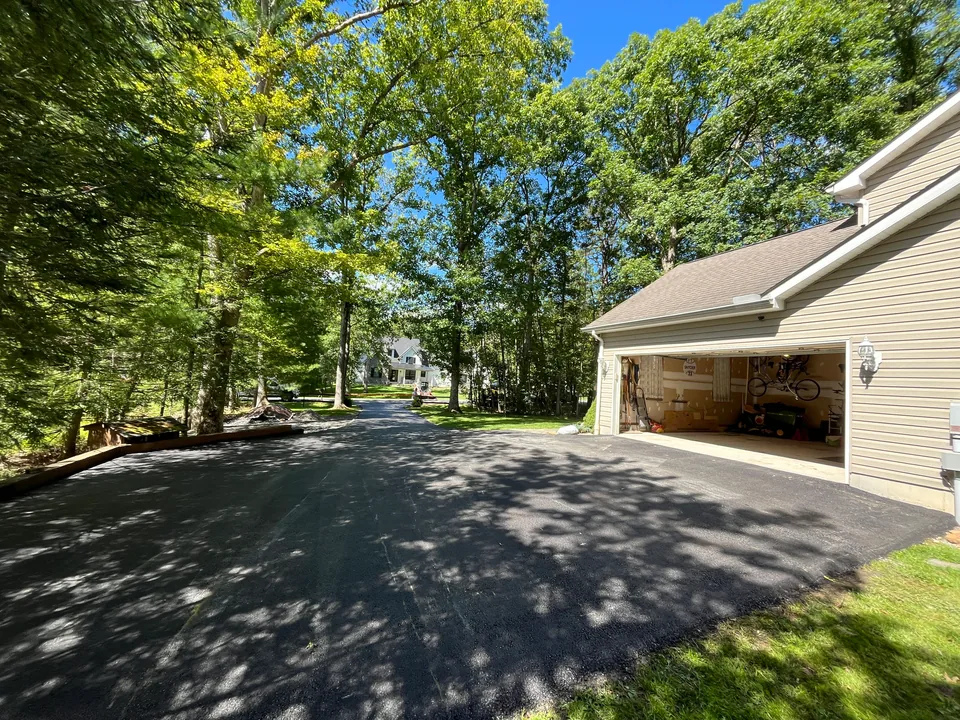 Freshly paved asphalt driveway beside an open two car garage under tall trees, asphalt driveway paving