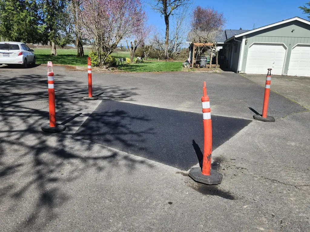 Finished rectangular hot asphalt patch on a residential driveway ringed by orange posts, asphalt patching and repair