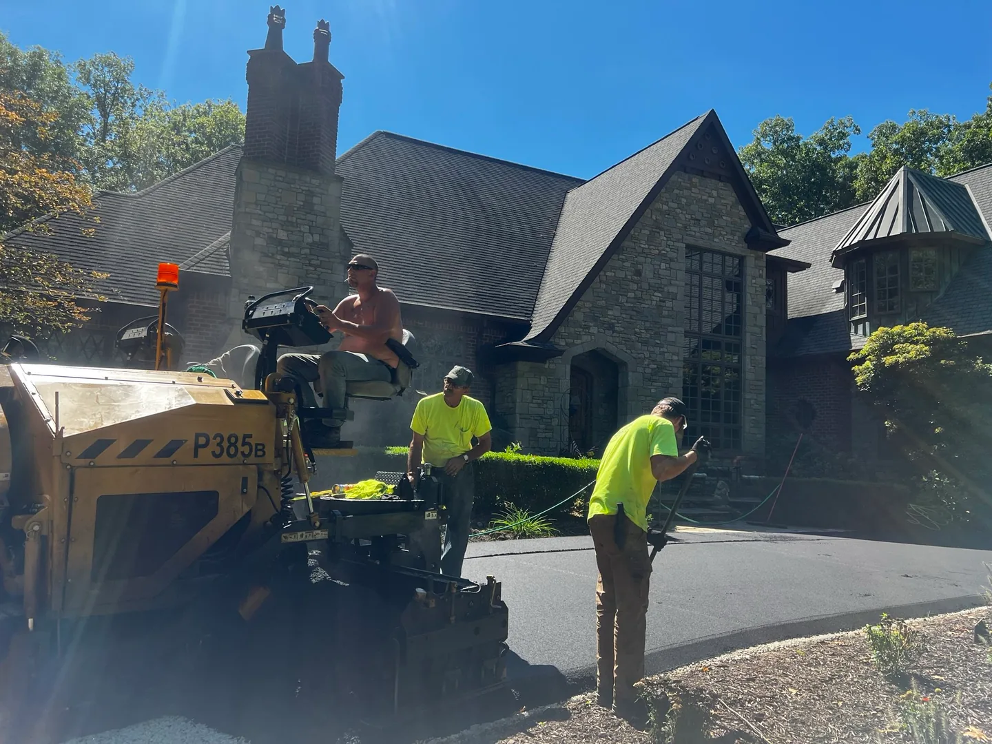Crew with a small paver laying a fresh asphalt layer on a driveway in front of a stone house, asphalt resurfacing