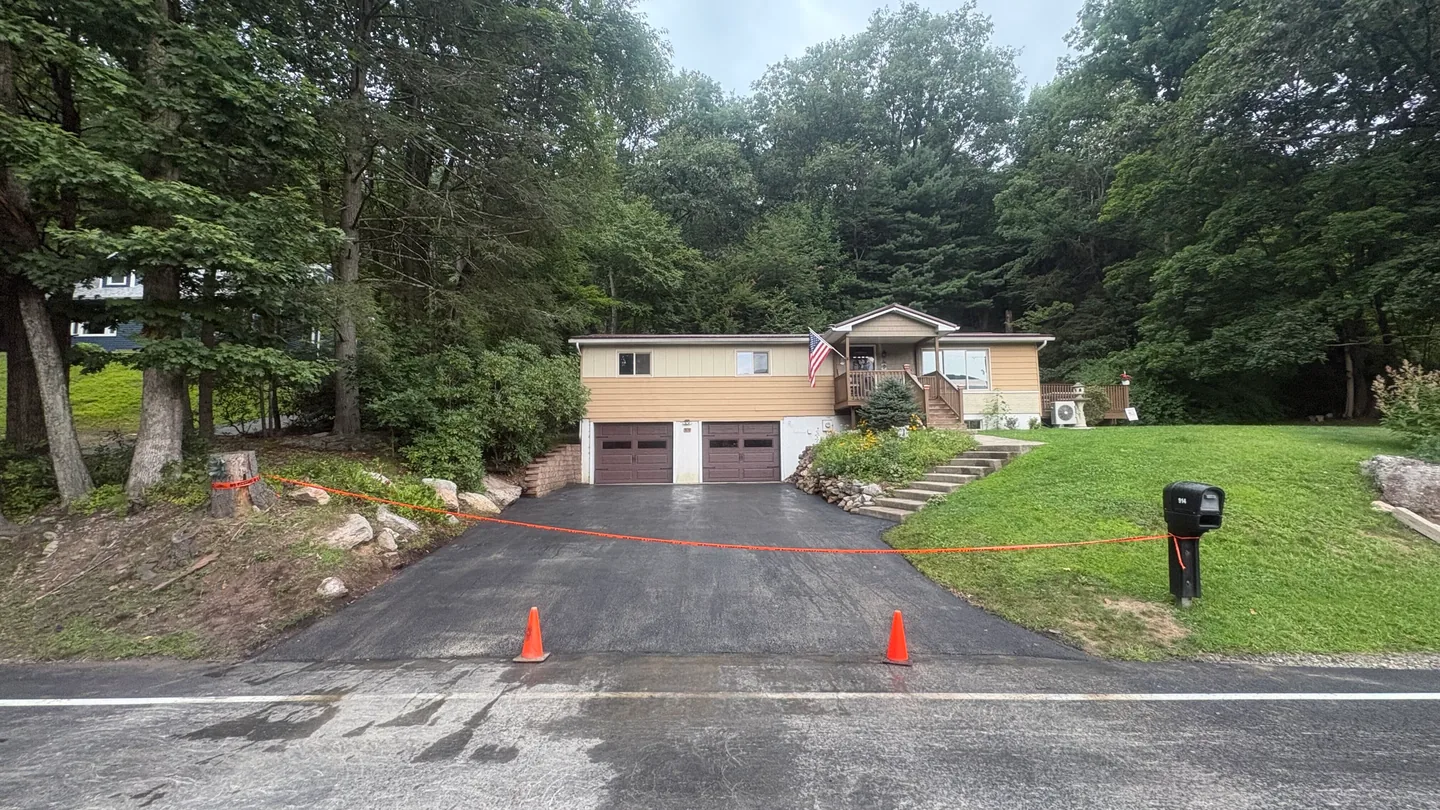 Freshly paved asphalt driveway with cones at the street below a raised ranch house, asphalt paving