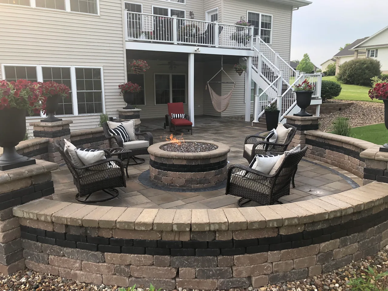 Paver patio with a round fire pit and a curved block seat wall behind a house, hardscaping and patios