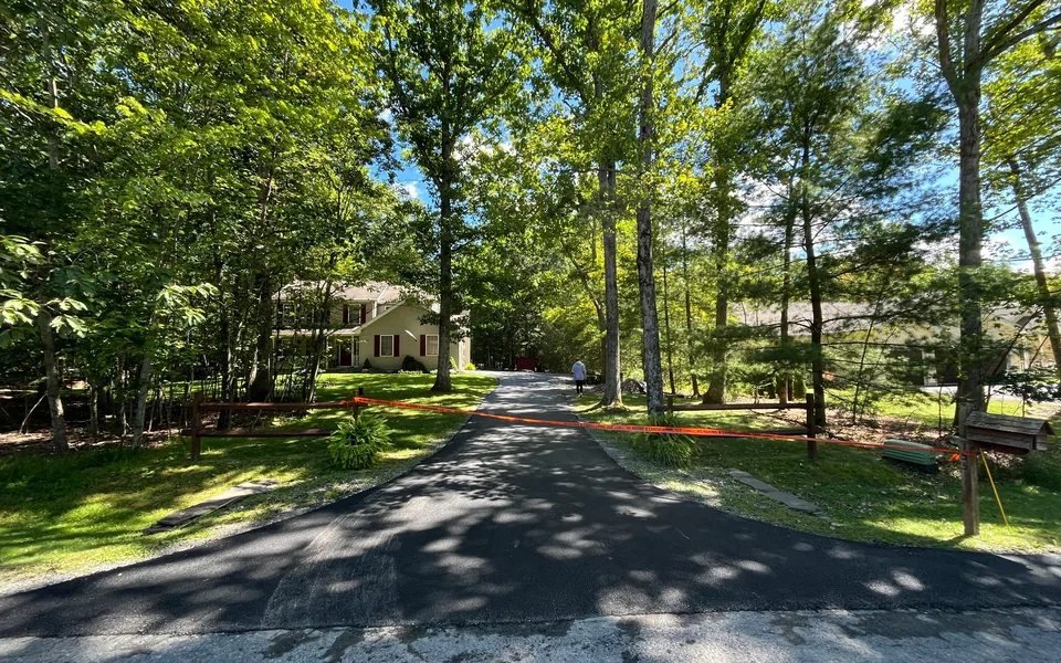 Long freshly paved asphalt drive through tall trees between split rail fences, private road paving