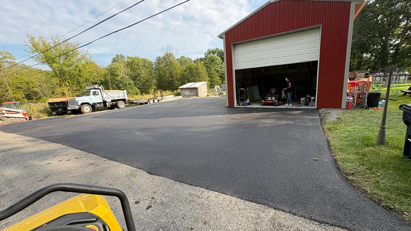 Fresh asphalt apron in front of a red pole barn with a dump truck and roller on site, asphalt paving and landscaping services